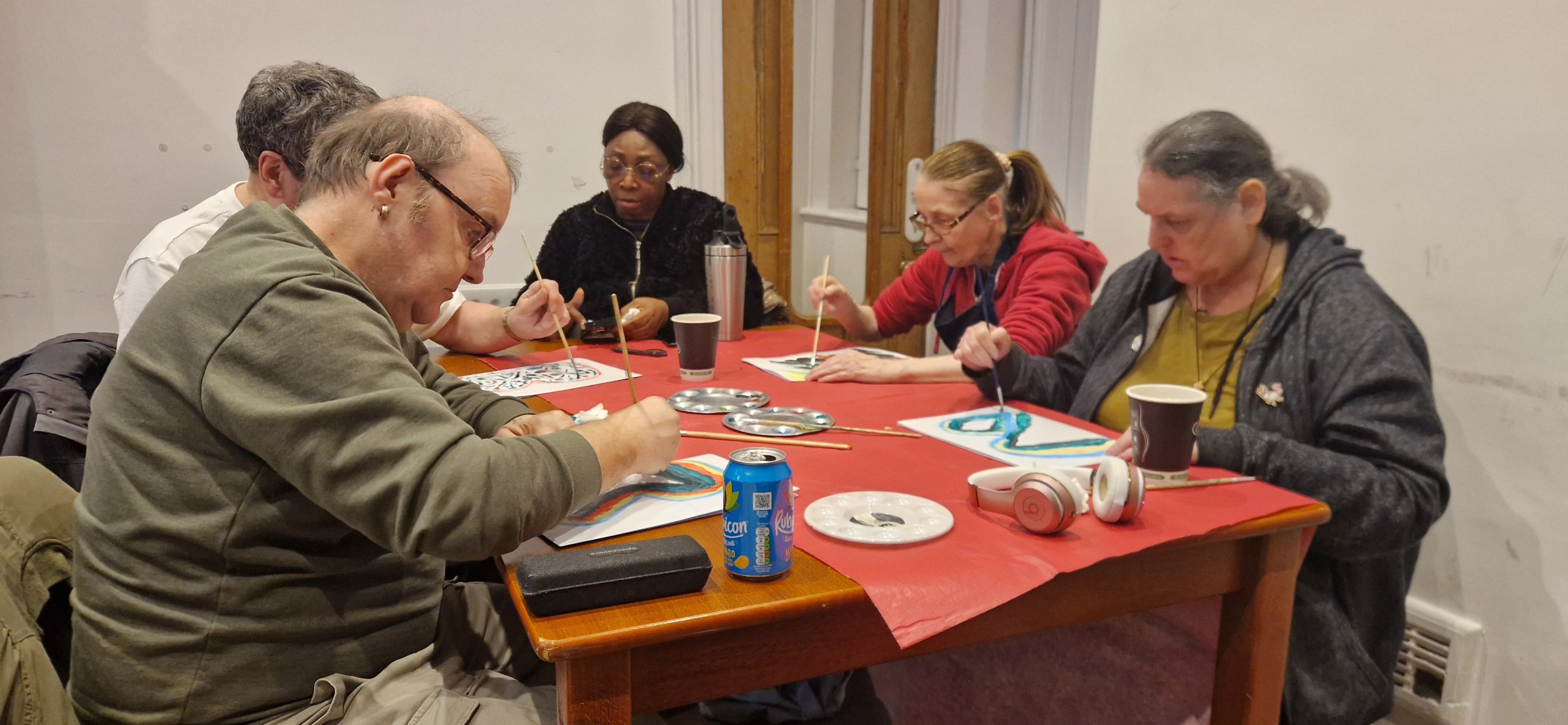 Group of adults painting together around a red table with brushes palettes and cups