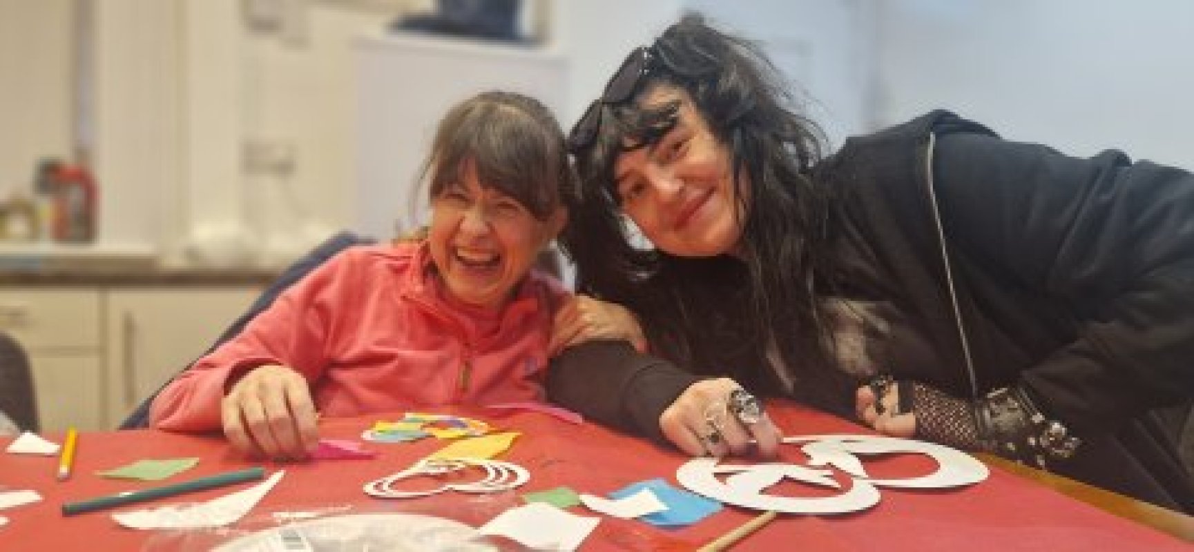 Two women smiling and leaning togetheer colourful craaterials at the table
