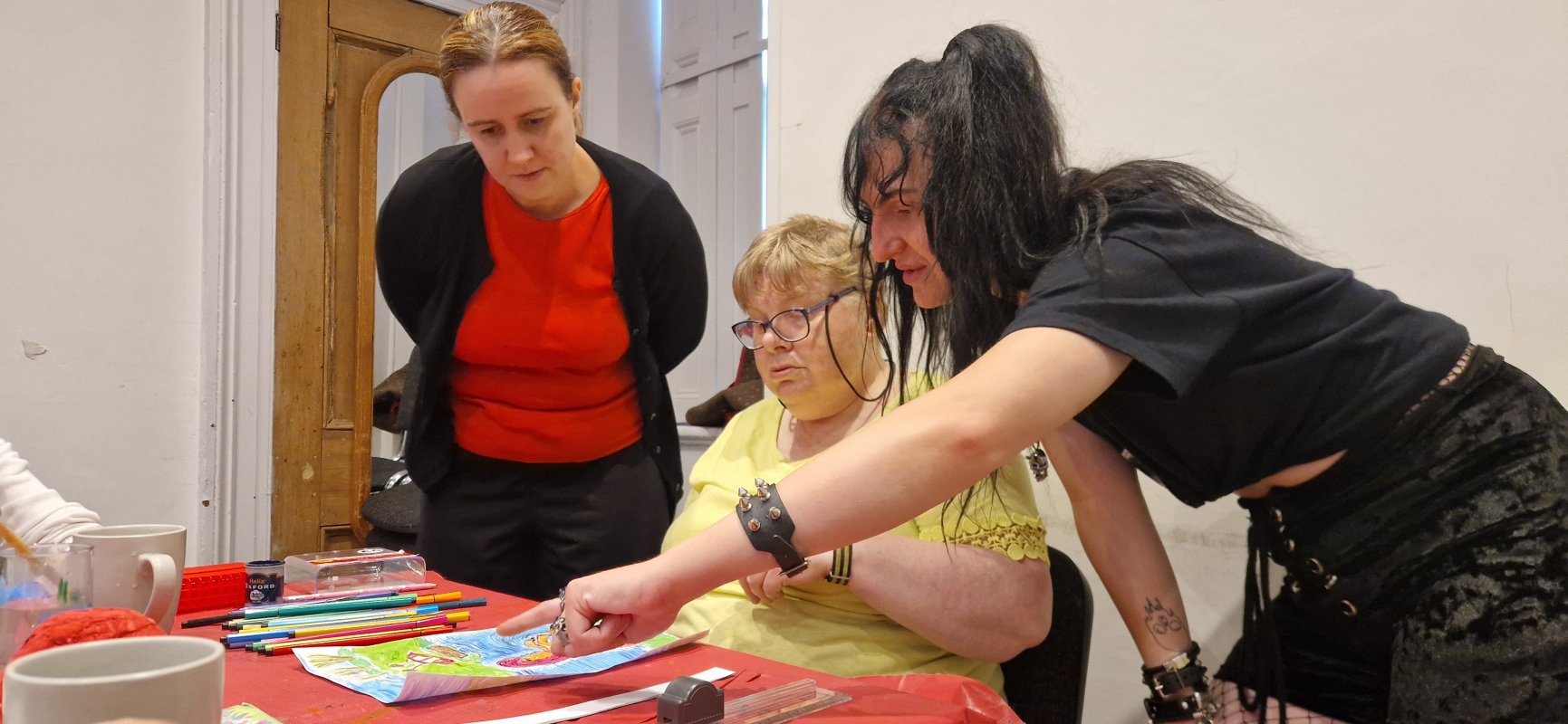 three people at a red table looking at some art worl